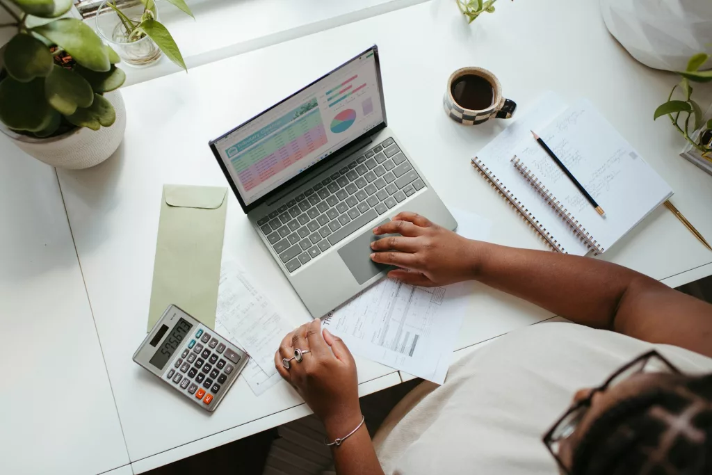 Eine Frau erstellt am Laptop mit Notizbuch und Taschenrechner einen Hochzeitsbudgetplan